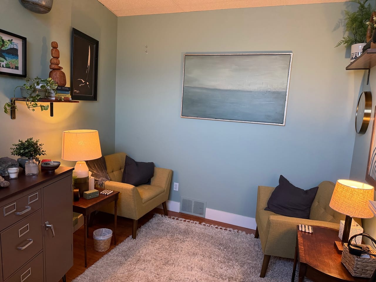 A sage-green therapy room with two mustard armchairs, warm table lamps and a calm painting of the sea