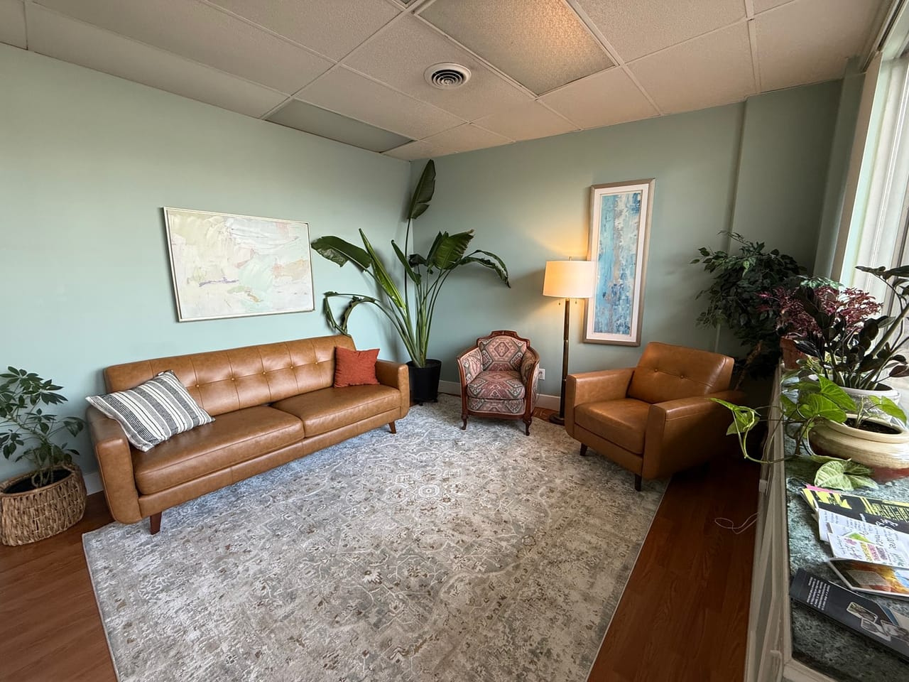 A bright waiting room with a tan leather sofa and armchairs, a patterned rug and plants along the window