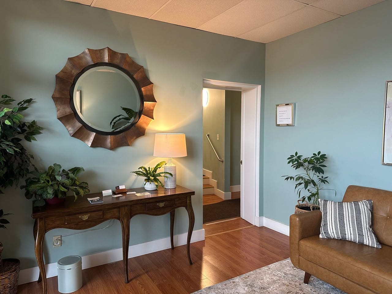 An entrance with a round wooden mirror over a console table, a lamp and a doorway to the stairs