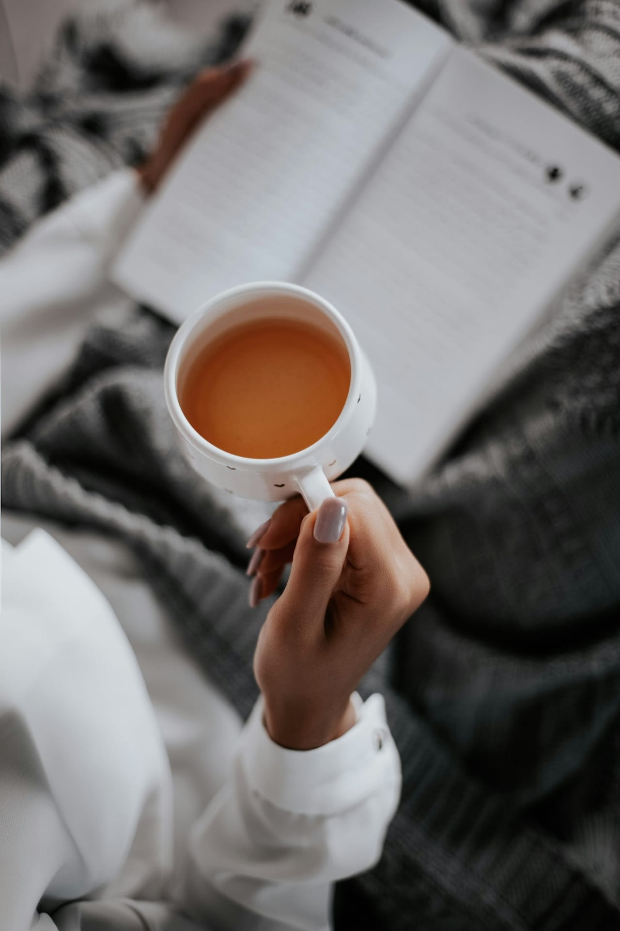 Hands holding a cup of tea over an open book and a knit blanket